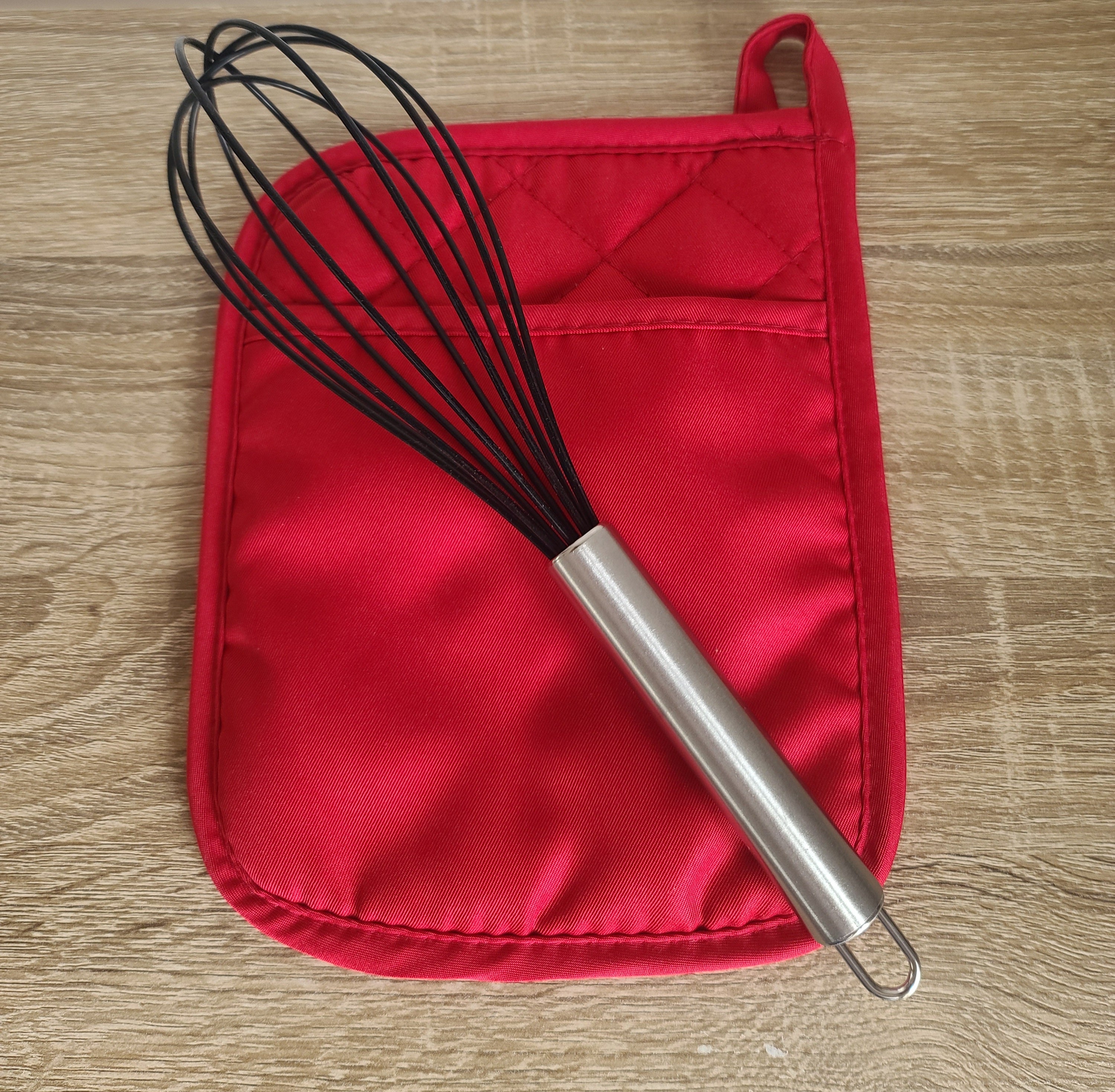 A kitchen whisk with a stainless steel handle and black wire loops, resting diagonally on a red quilted potholder over a light wood grain surface.
