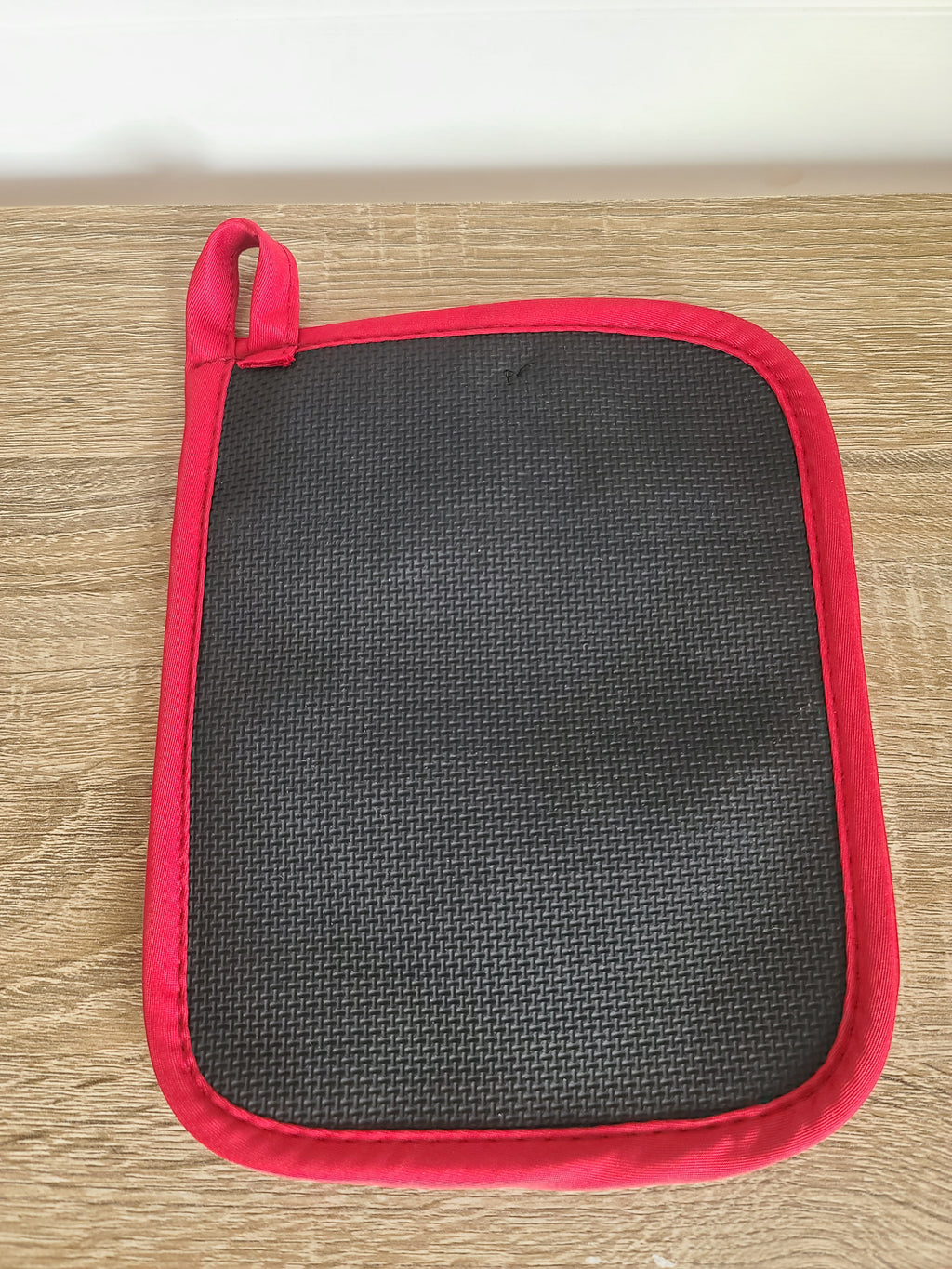 A rectangular potholder with a black textured center and a bright red border, featuring a matching red loop handle, shown on a light wood surface.