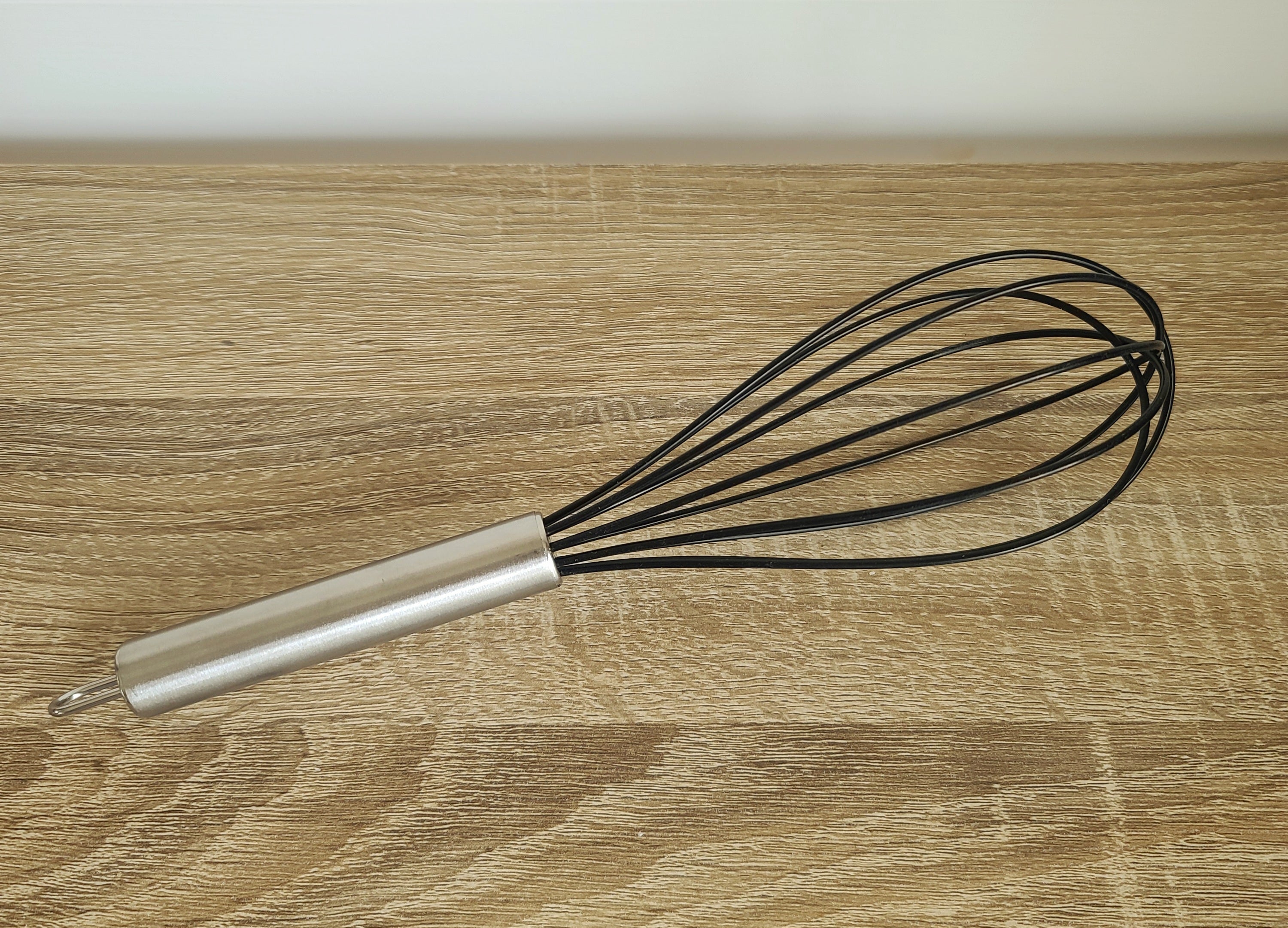 A metal whisk with a silver cylindrical handle and black wire loops, resting on a light brown wooden surface.